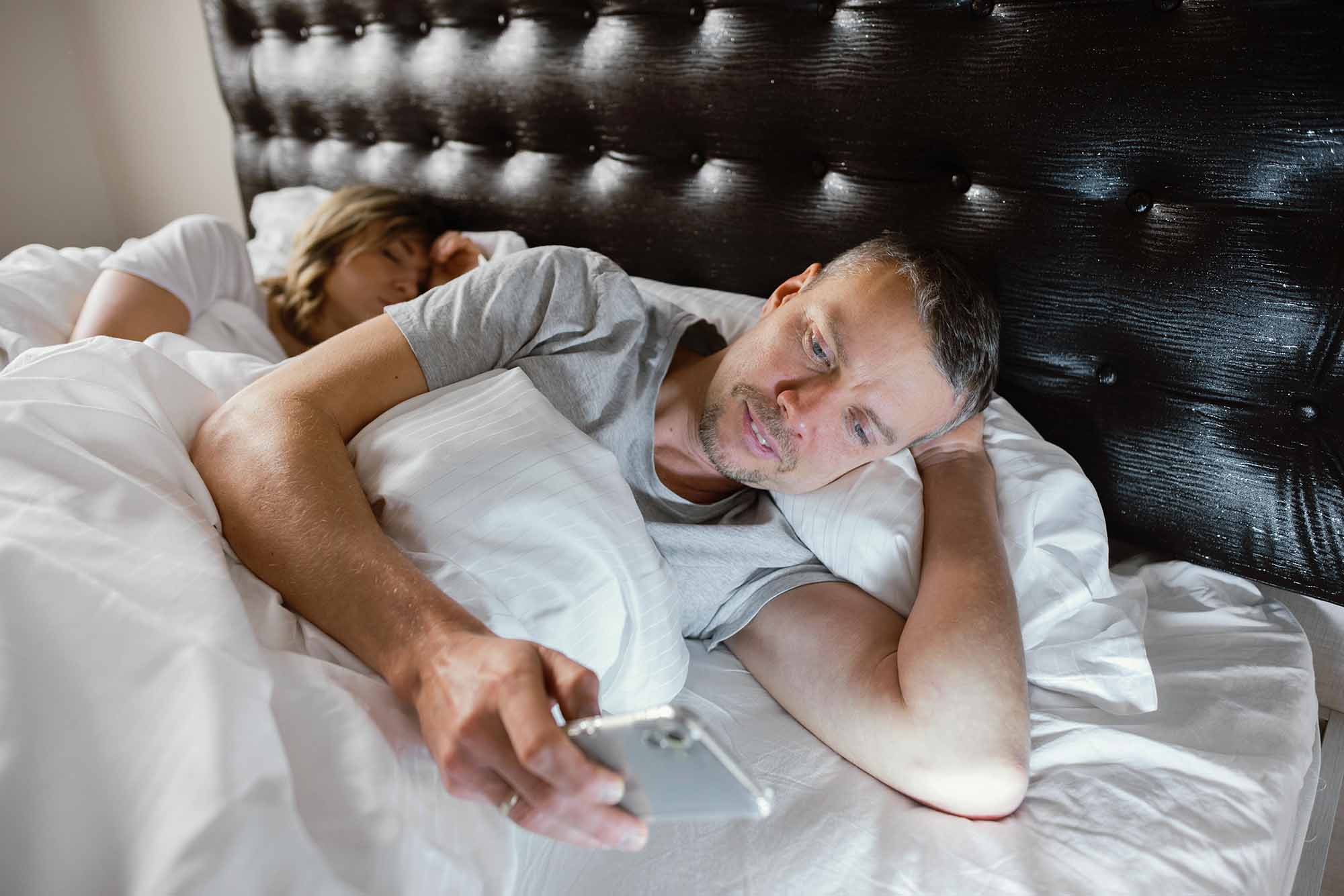Mann liegt wach im Bett und schaut auf sein Smartphone, während eine weitere Person im Hintergrund schläft.